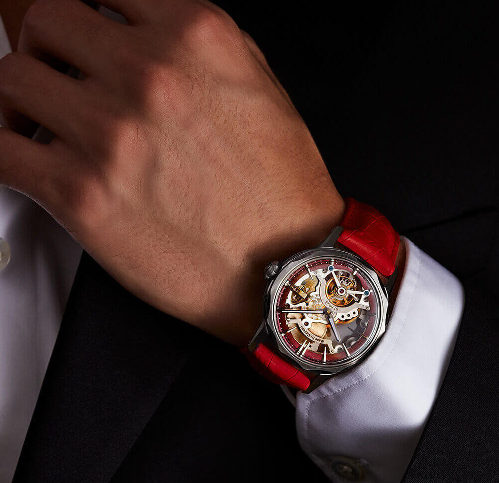 Close-up of a luxury skeleton tourbillon watch with a red leather strap worn with a tailored dark suit and white dress shirt.