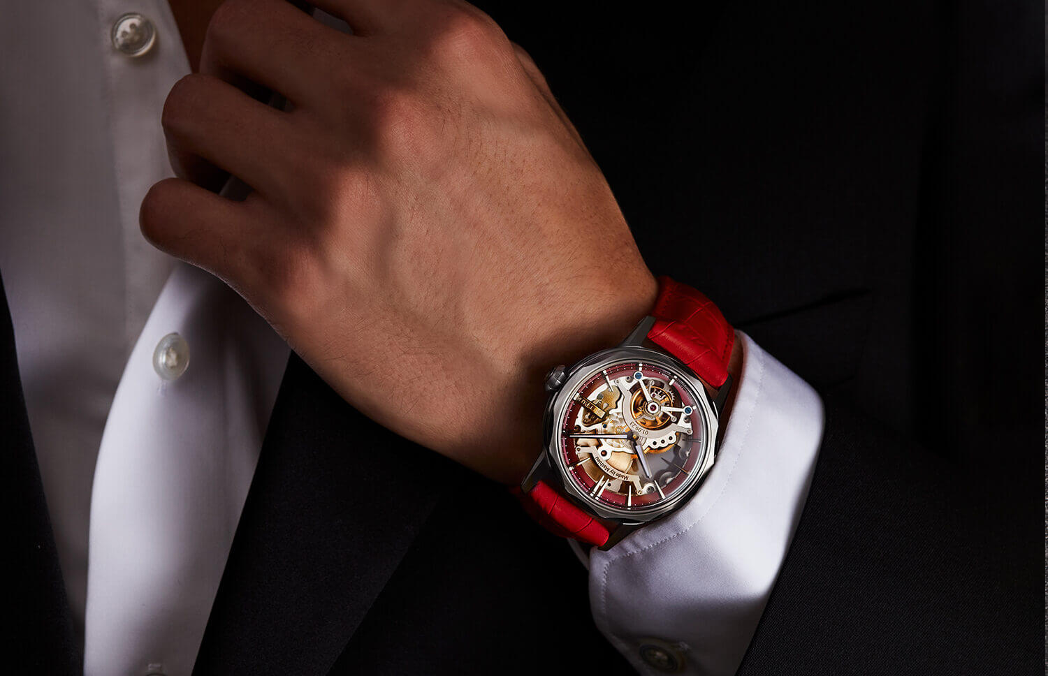 Close-up of a luxury skeleton tourbillon watch with a red leather strap worn with a tailored dark suit and white dress shirt.